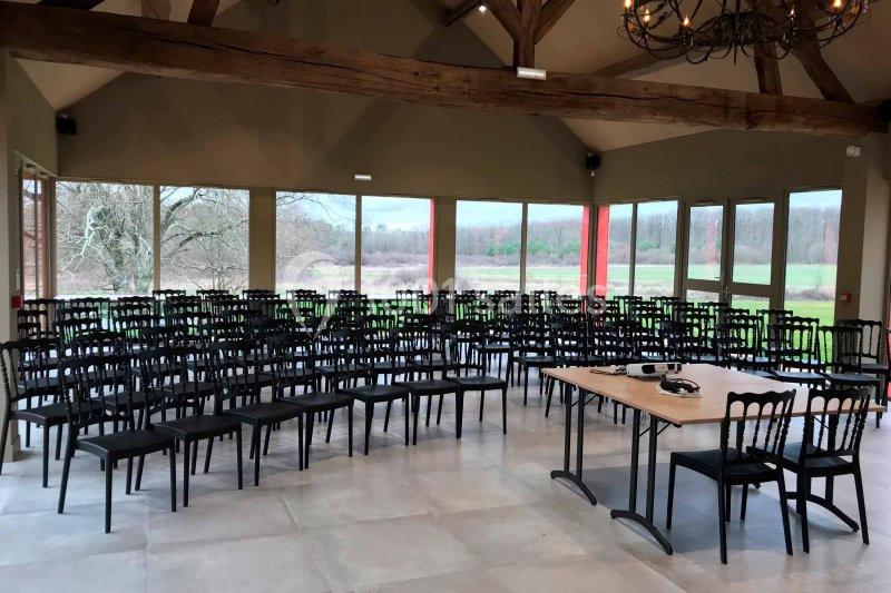 Salle lumineuse avec de grandes baies vitrées, rangées de chaises noires et une table équipée de matériel audio.
