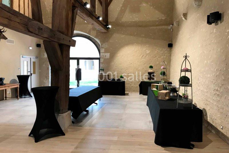 Salle avec poutres apparentes, tables noires dressées pour un événement et lumière naturelle entrant par une baie vitrée.