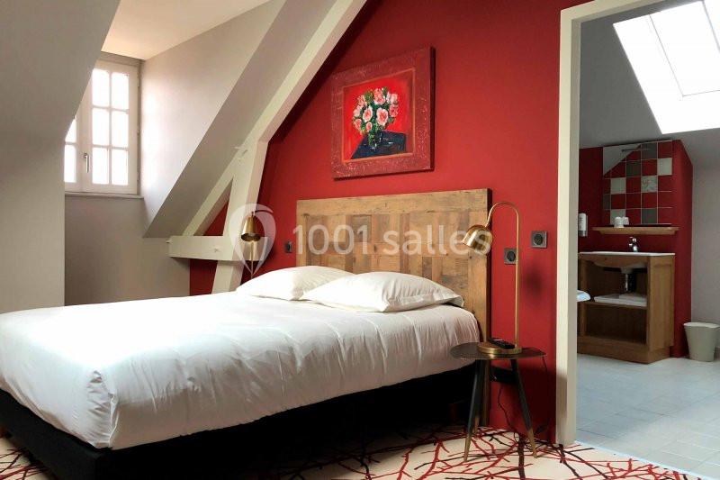 Chambre mansardée avec un lit double, mur rouge décoré d'un tableau floral, et salle de bain attenante visible.