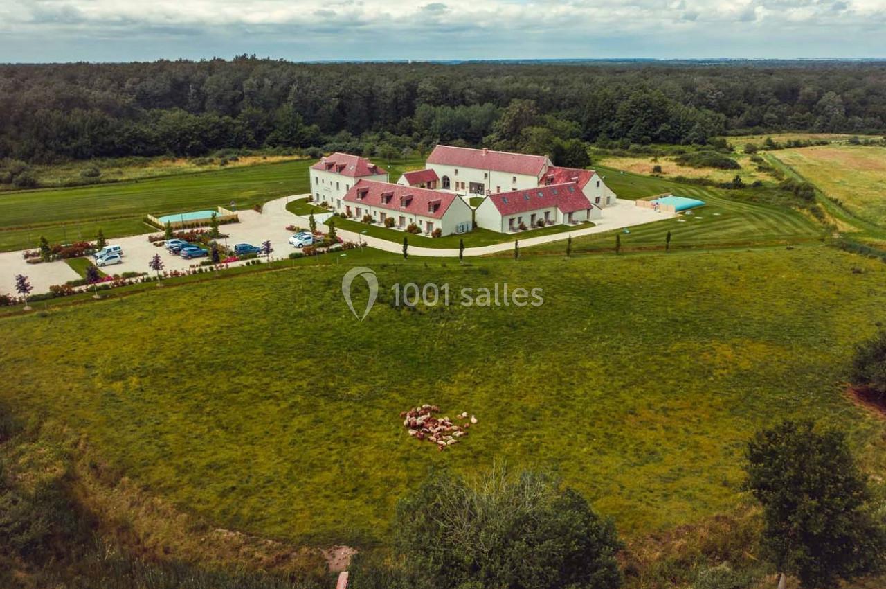 Vue aérienne d'un grand domaine avec bâtiments à toits rouges, piscine, parking et vastes espaces verts environnants.
