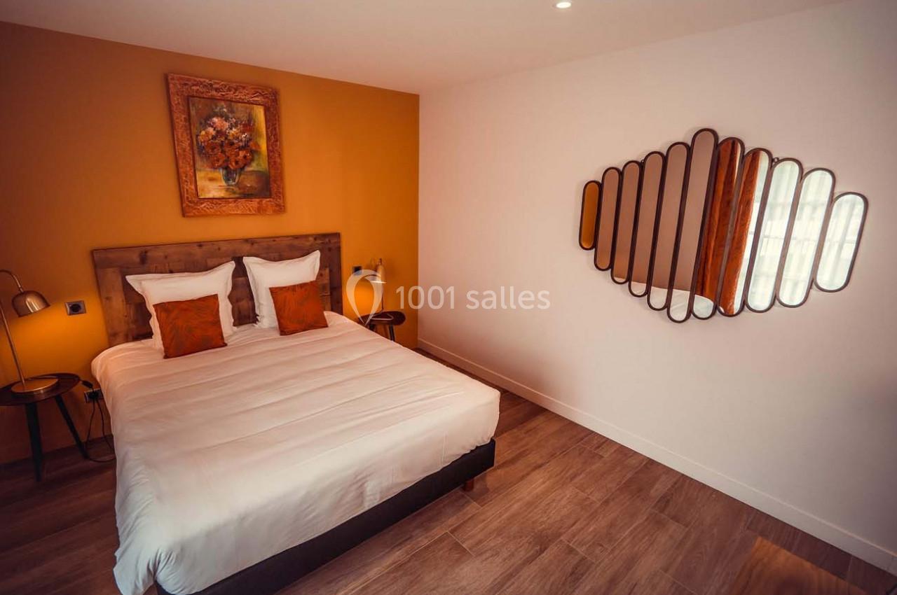 Chambre avec lit double, mur ocre, tableau floral, miroir design et sol en parquet.