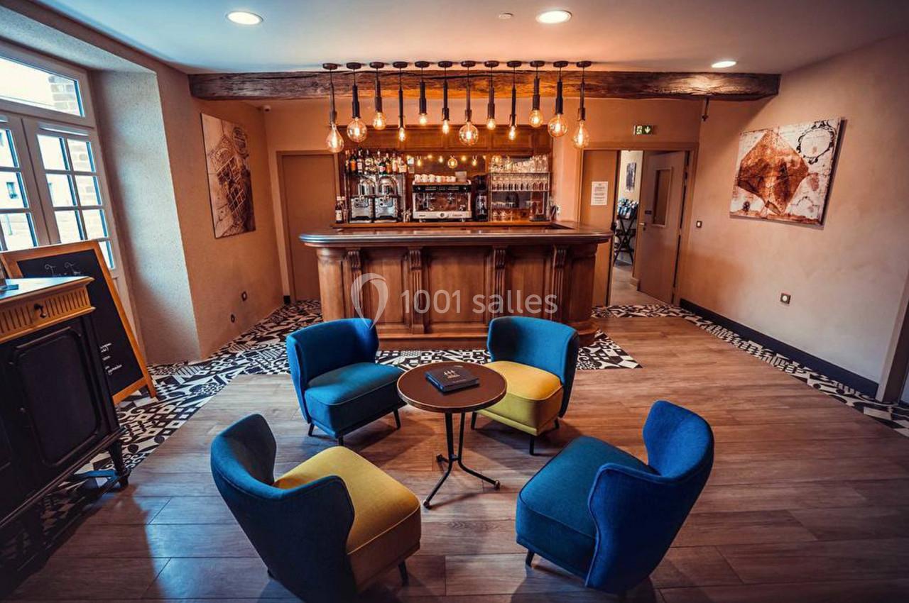 Espace lounge avec quatre fauteuils colorés autour d'une table basse, devant un bar en bois éclairé par des ampoules…