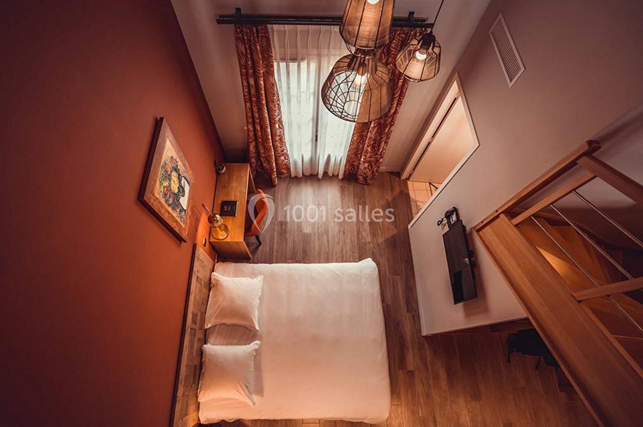 Vue aérienne d'une chambre avec lit double, parquet, bureau, rideaux rouges et éclairage suspendu.