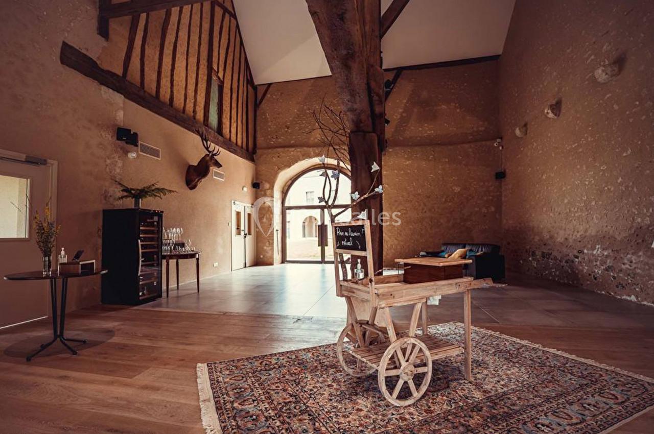 Salle spacieuse avec poutres apparentes, charrette en bois décorative au centre et mobilier sobre sur un tapis.