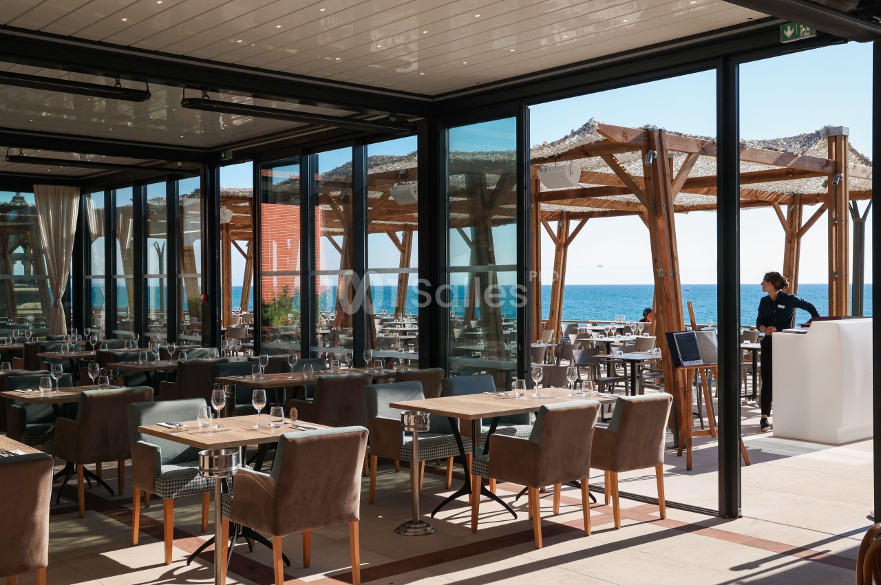 Salle de restaurant vitrée aux tables dressées de verres, face à une terrasse en bois ouverte sur la mer.
