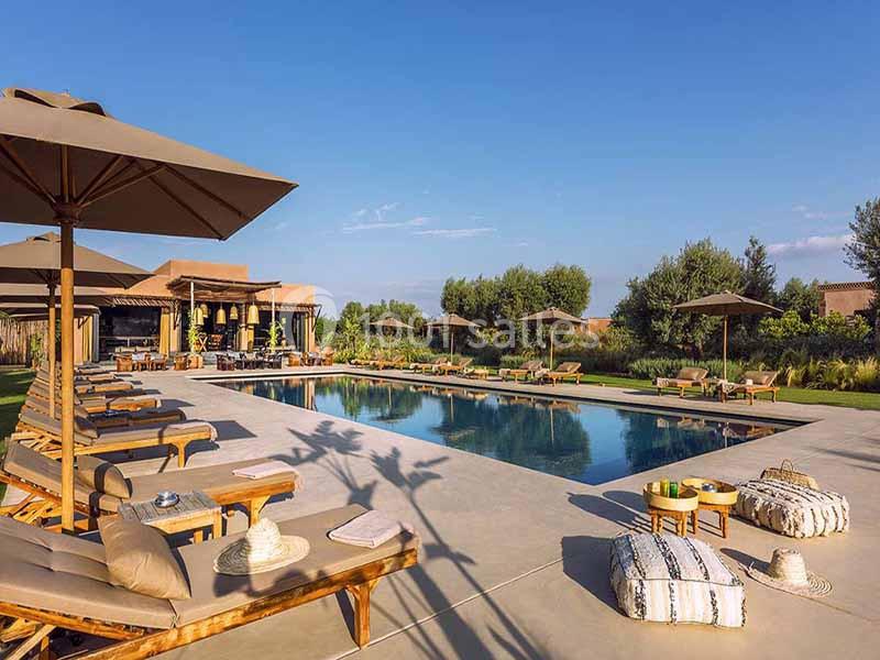 Piscine extérieure entourée de chaises longues, parasols et coussins, avec jardin et bâtiment en arrière-plan.