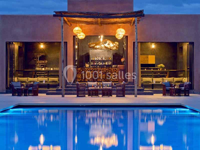 Vue d'une terrasse éclairée avec un bar central, des tables et une piscine au premier plan, en soirée.