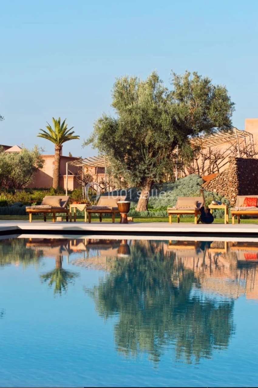 Piscine calme entourée de végétation méditerranéenne, avec des chaises longues et un ciel dégagé en arrière-plan.