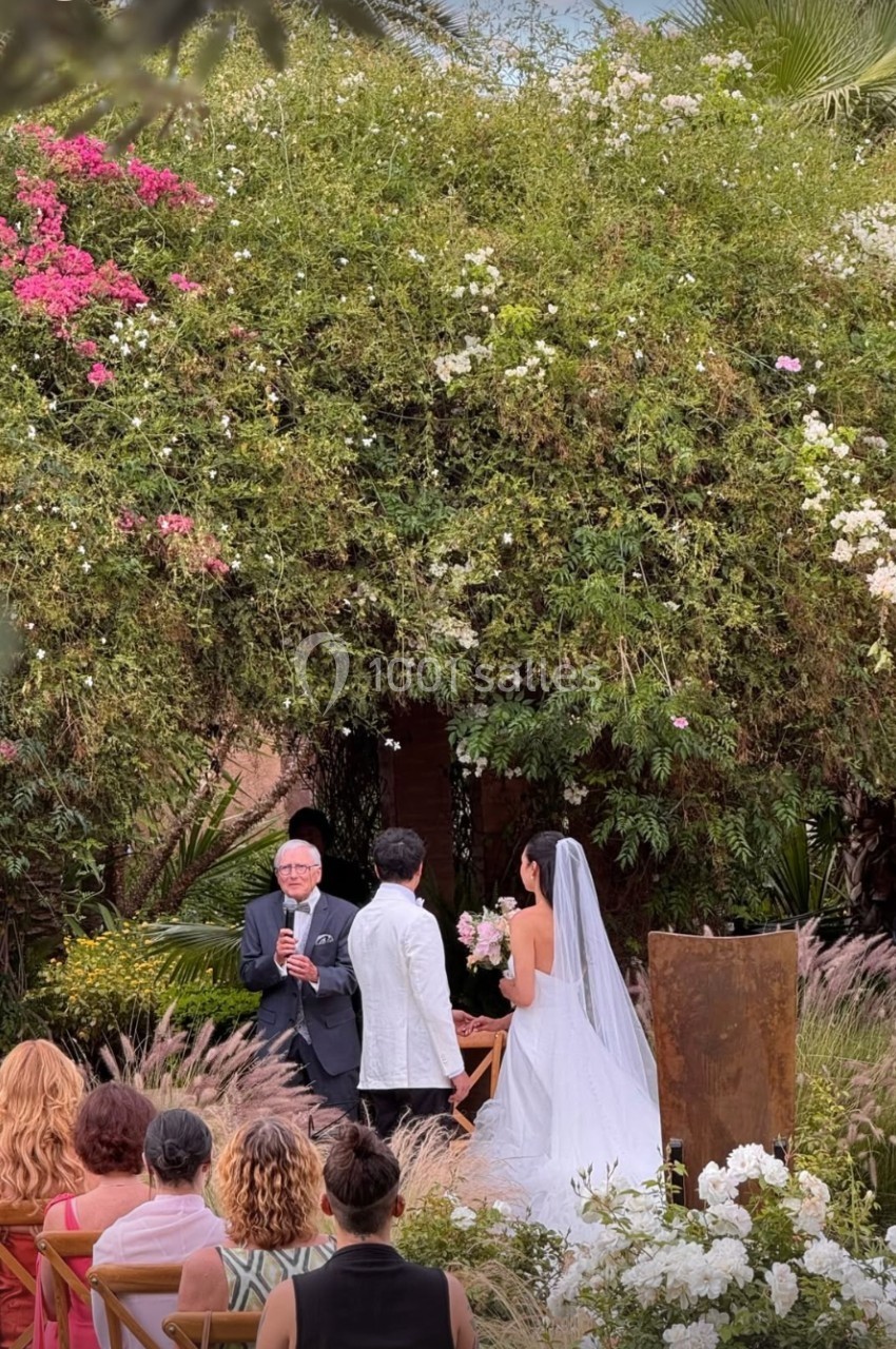 Un couple se marie en plein air devant un officiant, entouré de végétation luxuriante et d'invités assis.