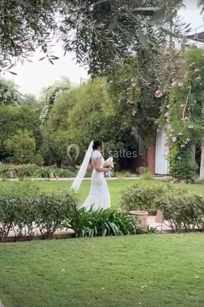 Une mariée en robe blanche marche dans un jardin verdoyant, tenant un bouquet de fleurs.