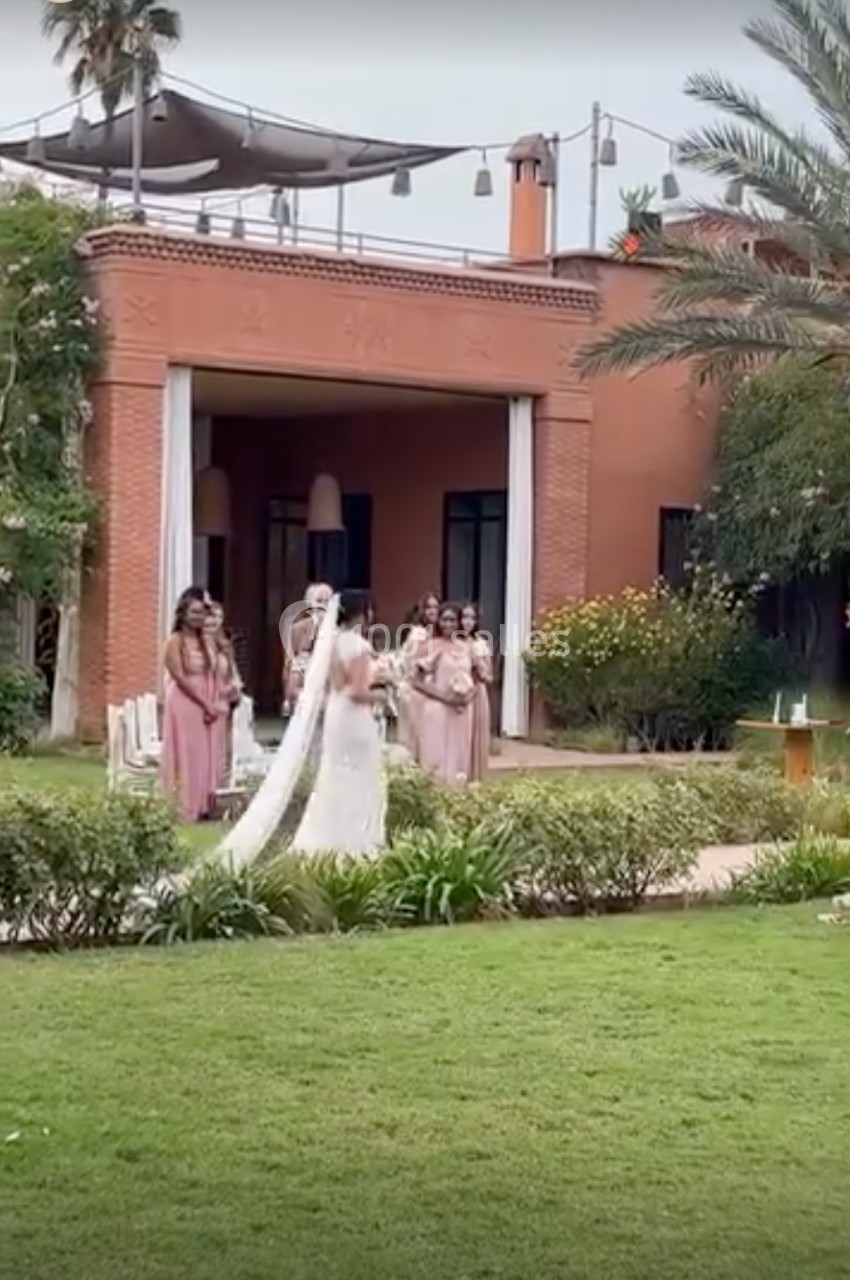 Une mariée en robe blanche marche dans un jardin verdoyant, suivie par des demoiselles d'honneur en robes roses.
