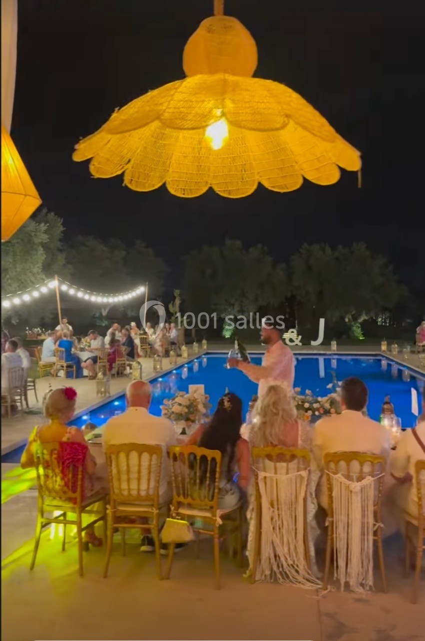 Des convives dînent en extérieur près d'une piscine éclairée, sous des lampes suspendues, en soirée.