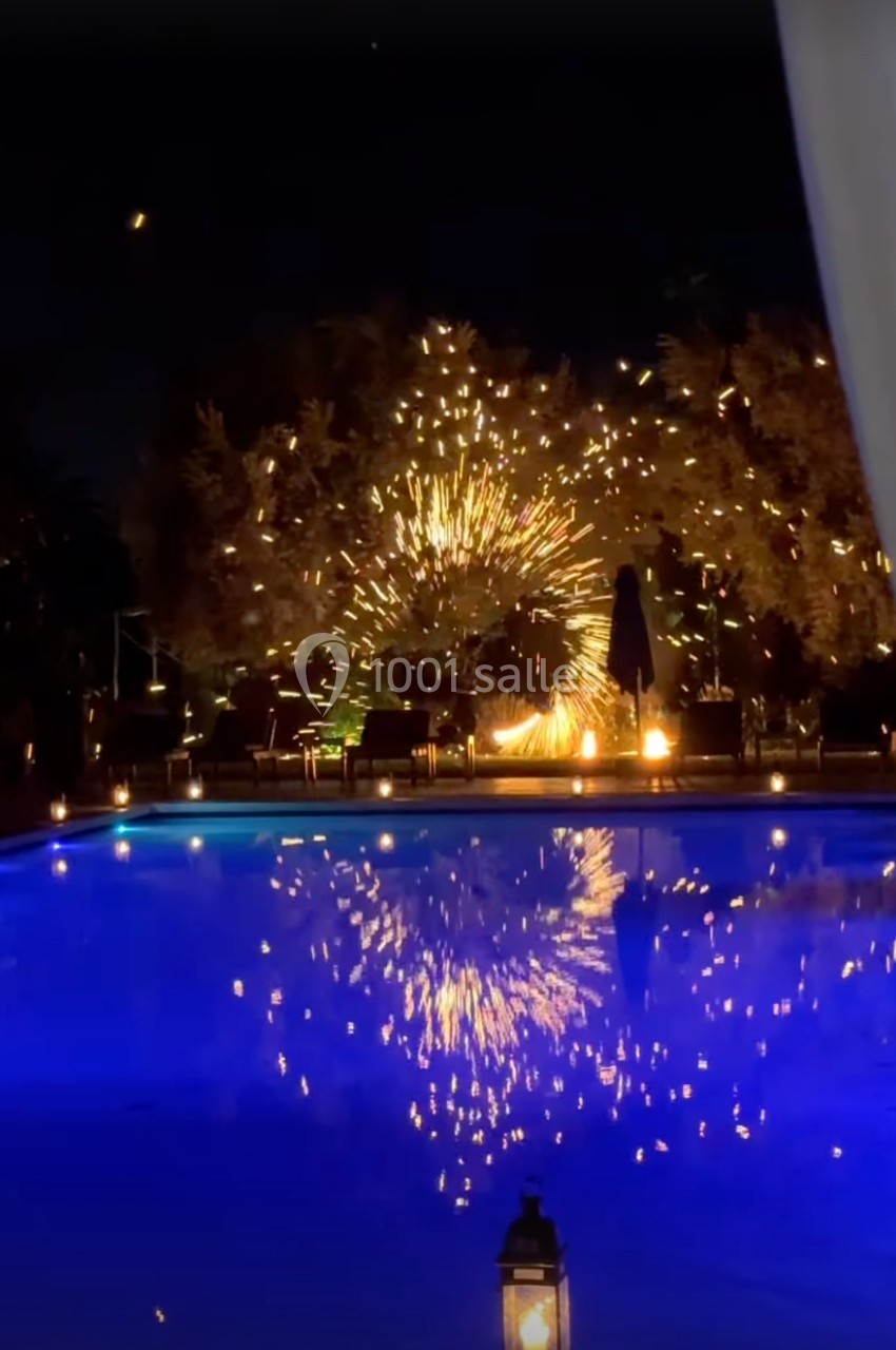 Feu d'artifice illuminant un jardin avec piscine de nuit, reflété dans l'eau calme.