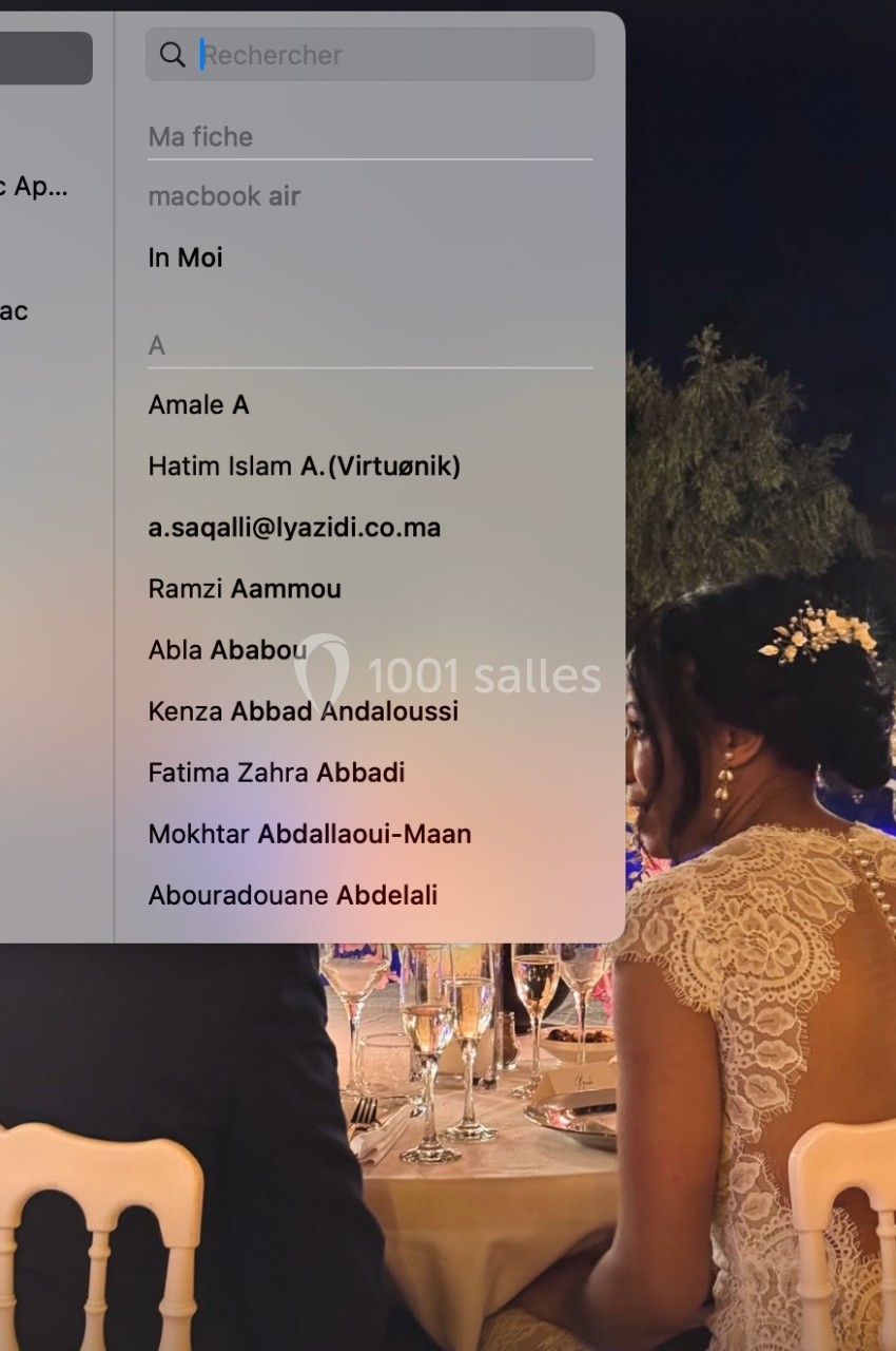 Une femme en robe de mariée assise à une table lors d'une réception en soirée, avec un écran d'ordinateur au premier plan.