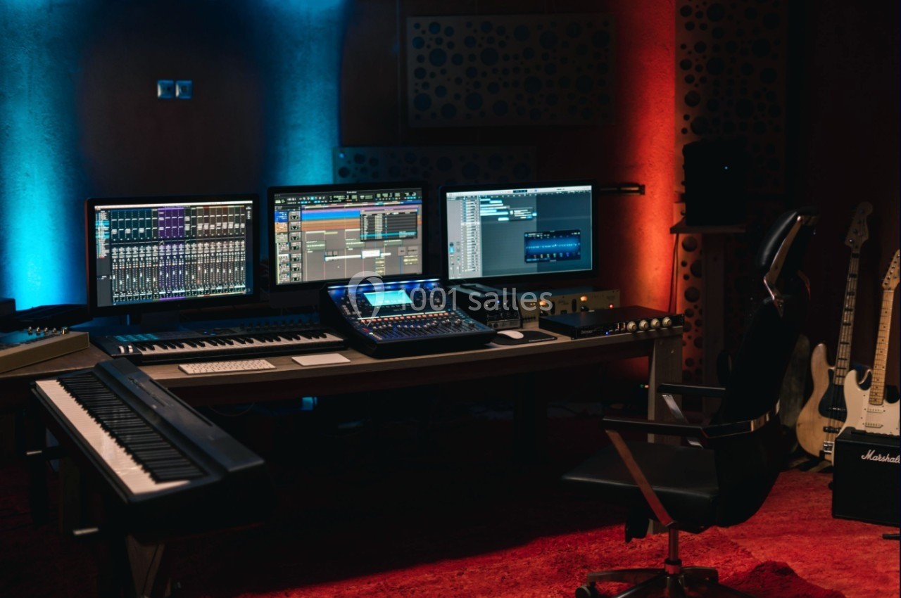 Poste de travail de studio d'enregistrement avec écrans, clavier, console audio et guitares dans une ambiance éclairée.