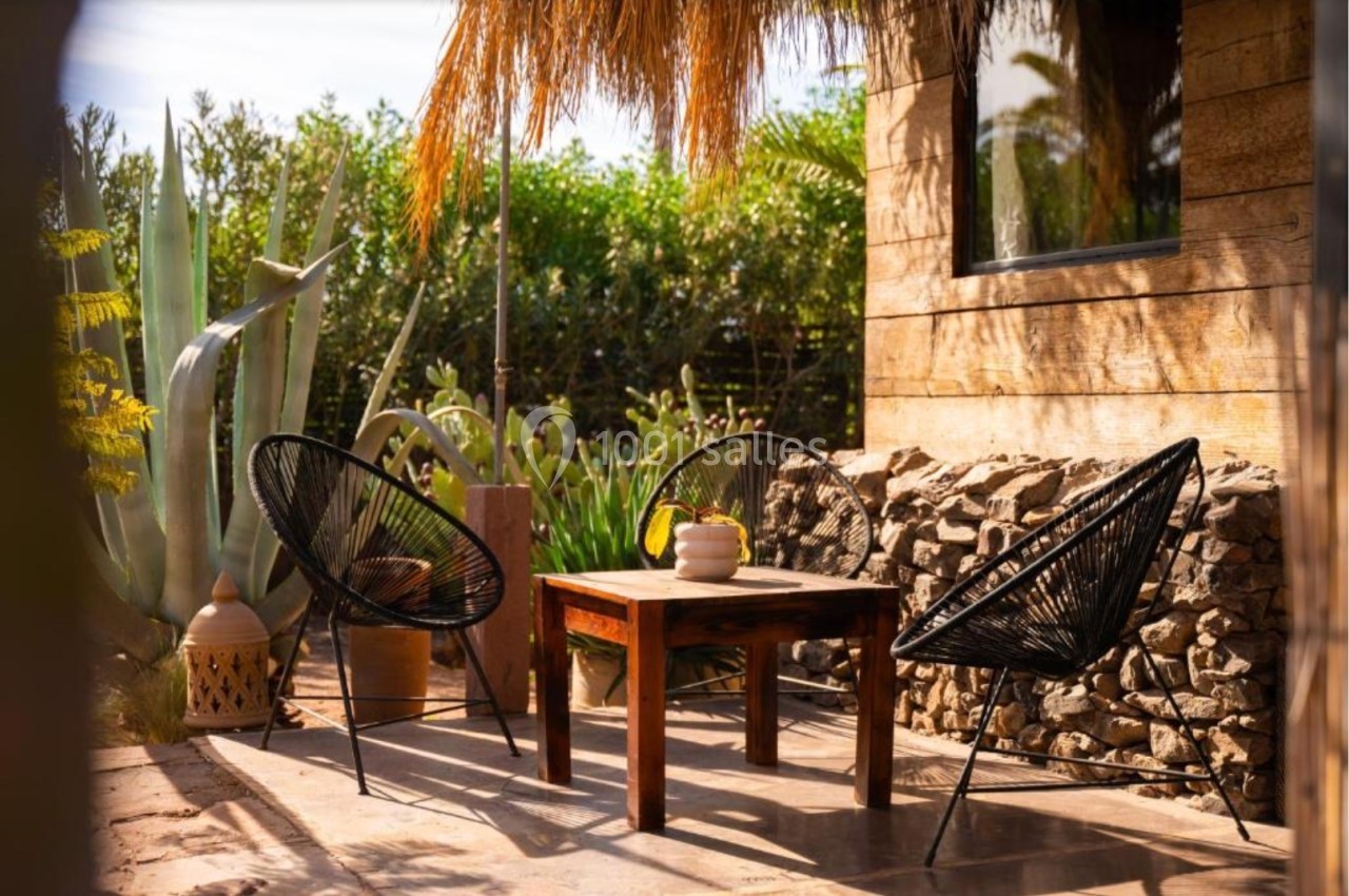 Terrasse ensoleillée avec table en bois, deux chaises noires et plantes luxuriantes en arrière-plan.