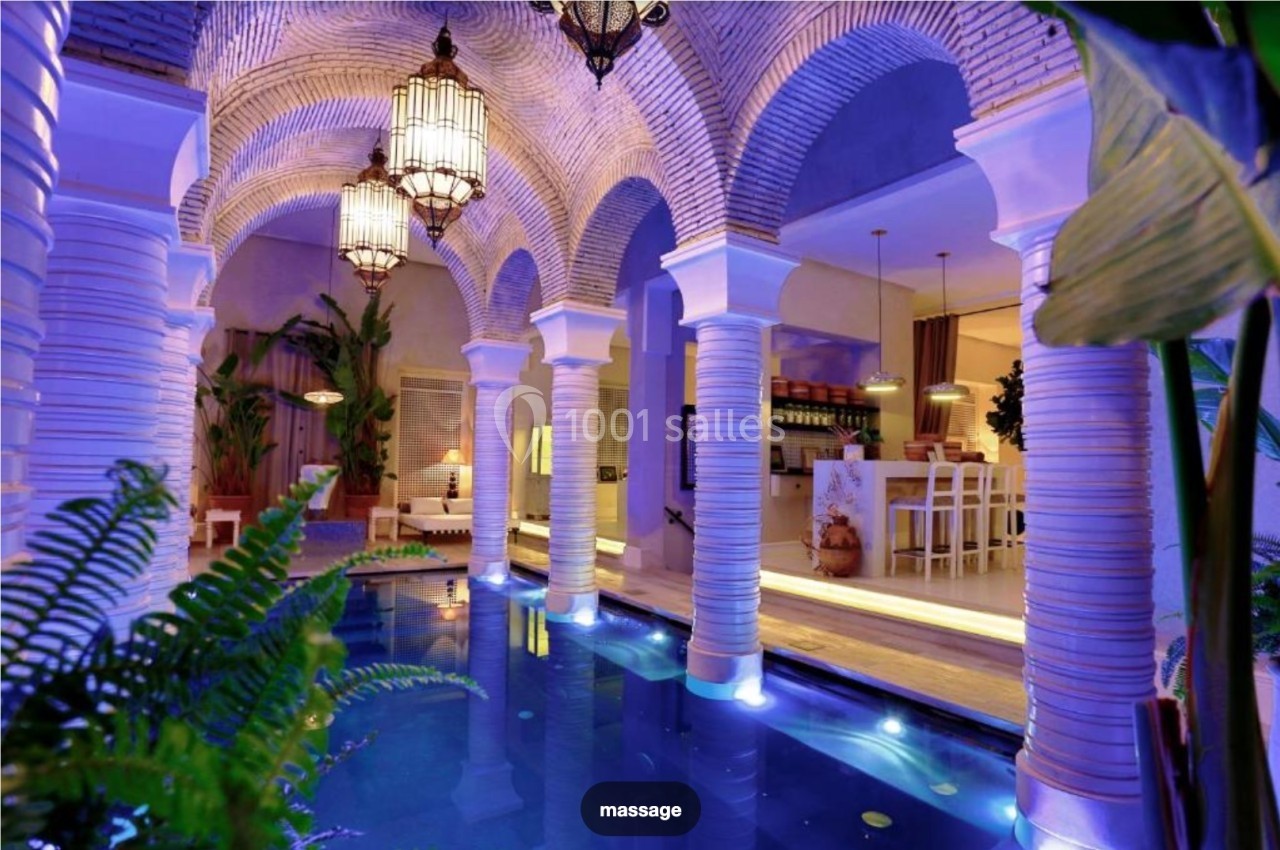 Intérieur d'un riad avec piscine éclairée, arches en pierre, luminaires suspendus et plantes tropicales.