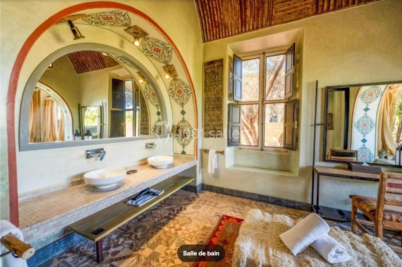 Salle de bain lumineuse avec double vasque, miroirs encadrés d'arcs décorés, fenêtre ouverte et tapis au sol.