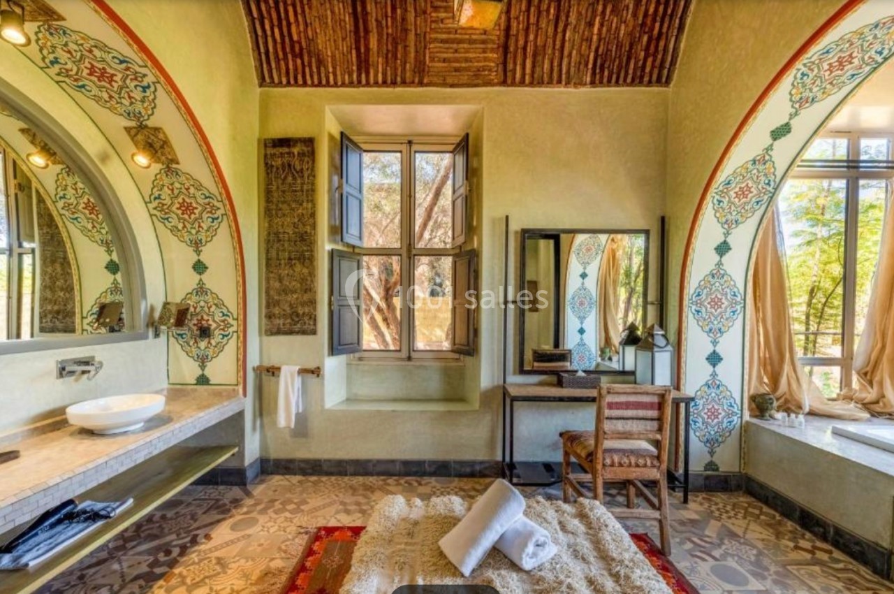 Salle de bain lumineuse avec décorations orientales, lavabo, miroir, baignoire, tapis et vue sur un jardin par une fenêtre.