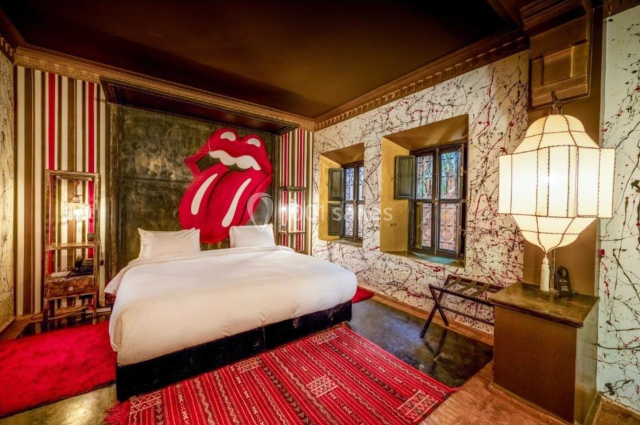 Chambre avec lit double, décoration murale artistique, tapis rouge et grandes fenêtres laissant entrer la lumière naturelle.