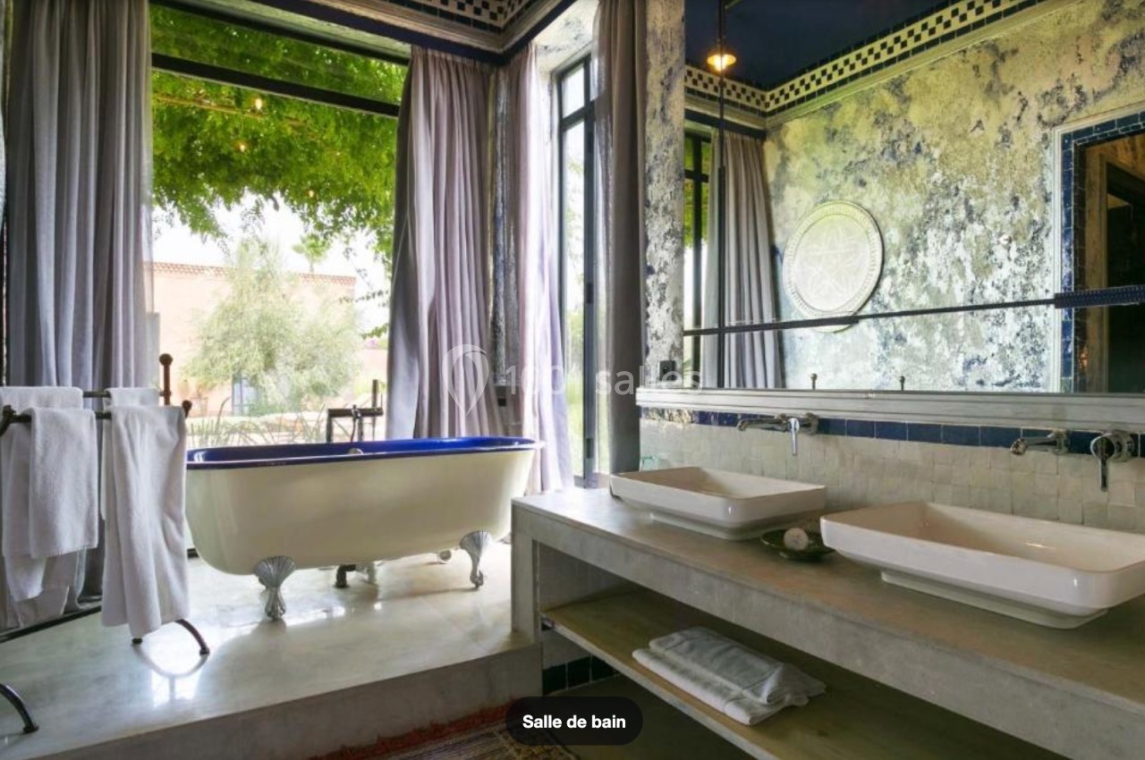 Salle de bain lumineuse avec baignoire sur pieds, double vasque, miroirs et grandes fenêtres donnant sur un jardin.