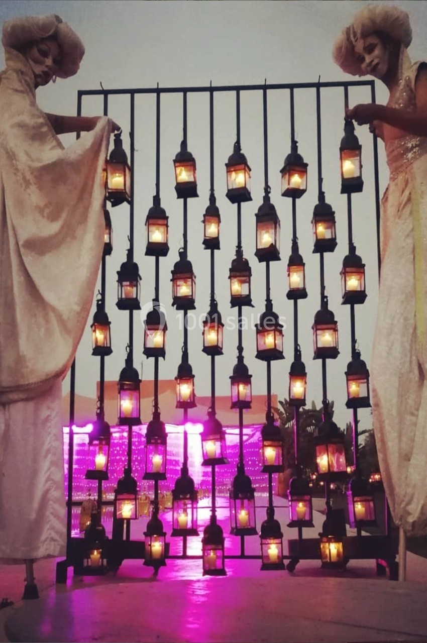 Deux personnes sur échasses en costumes blancs, tenant une structure décorée de lanternes allumées au crépuscule.