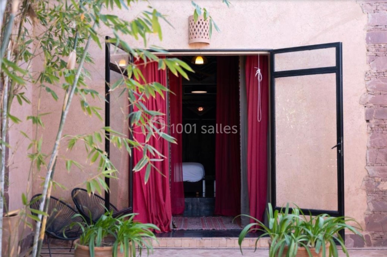 Porte ouverte sur une pièce avec rideaux rouges, vue sur un lit, entourée de plantes et de bambous.