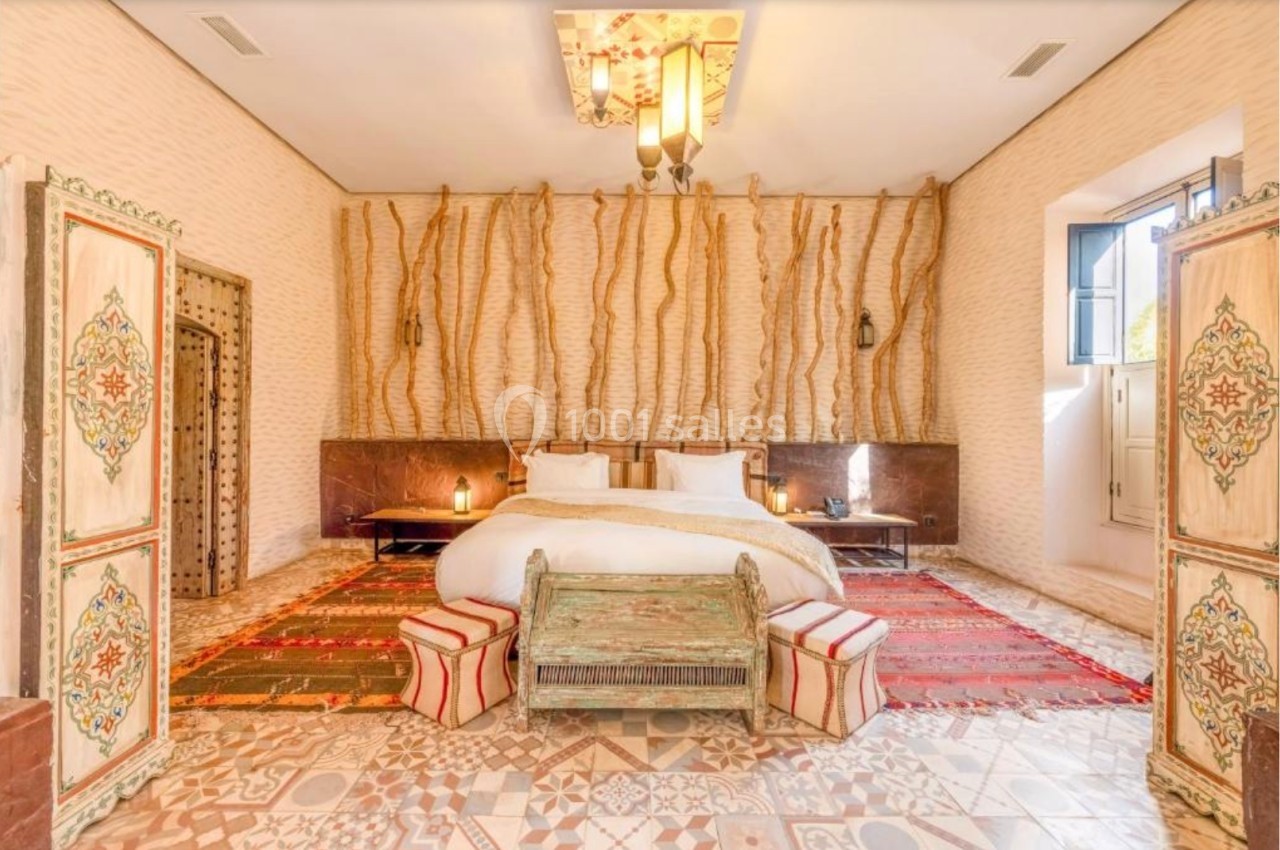 Chambre lumineuse avec lit double, tapis colorés, murs texturés et décorations traditionnelles en bois sculpté.