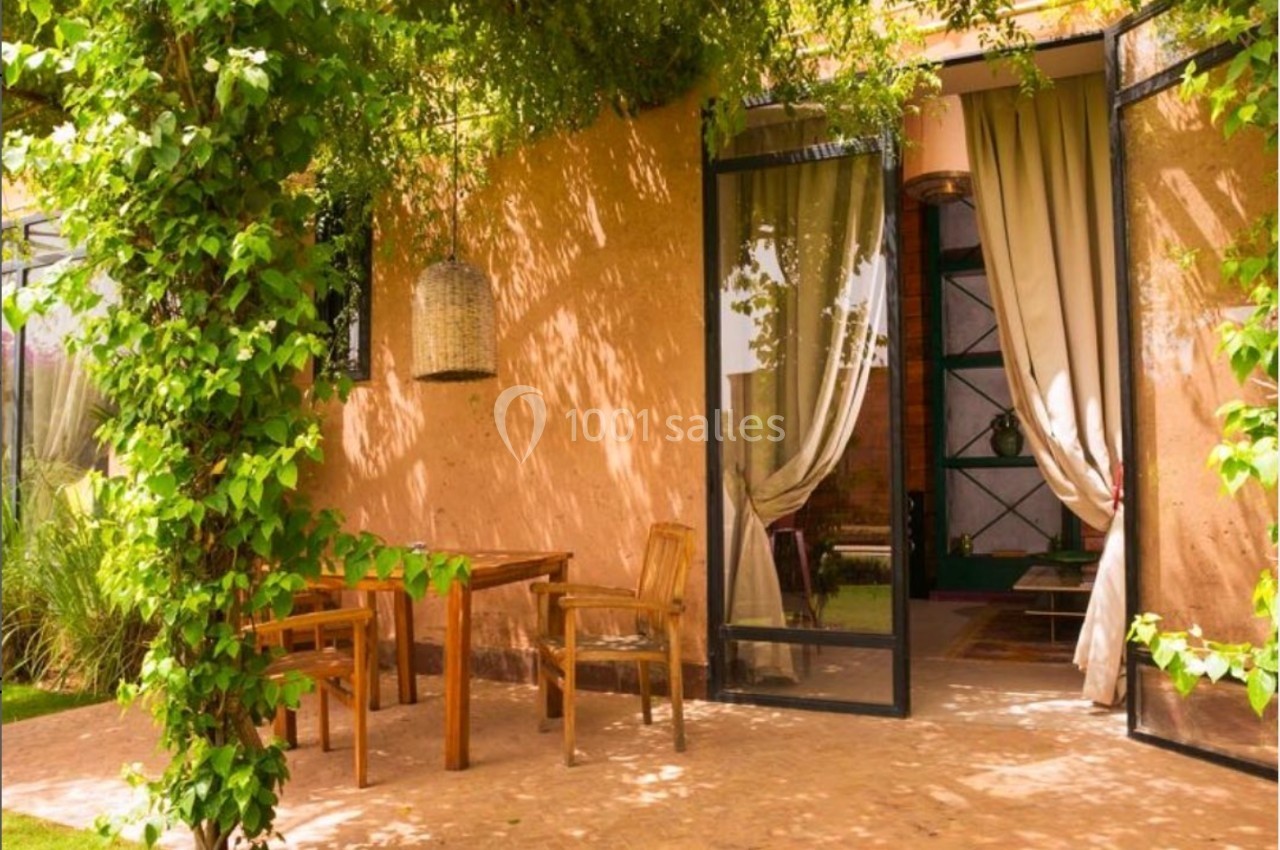 Terrasse ombragée avec table et chaises en bois, entourée de verdure, donnant sur une maison aux murs couleur terre.