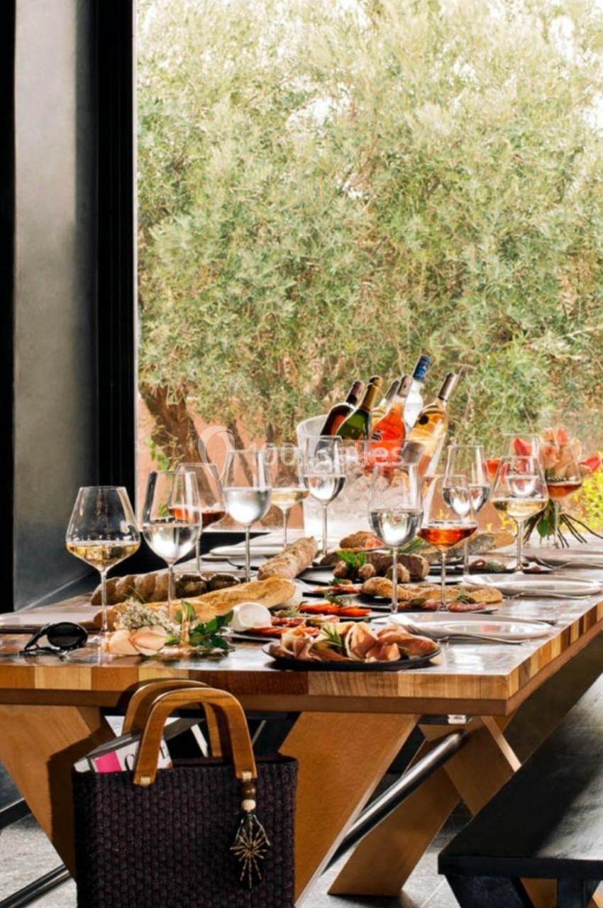 Table en bois dressée avec des plats variés, des verres à vin et des bouteilles, devant une grande fenêtre et des oliviers.