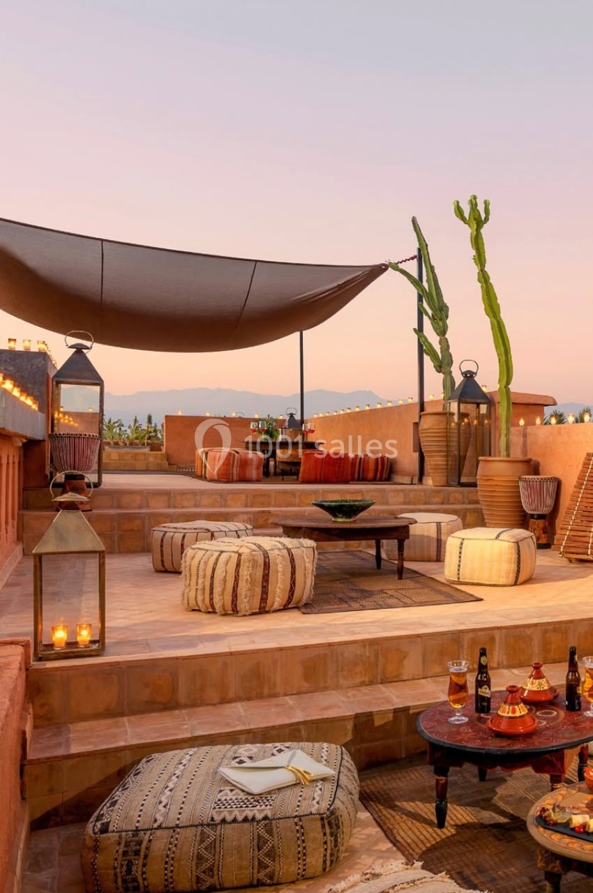 Terrasse aménagée avec coussins, tables basses, lanternes et vue sur un paysage au coucher du soleil.