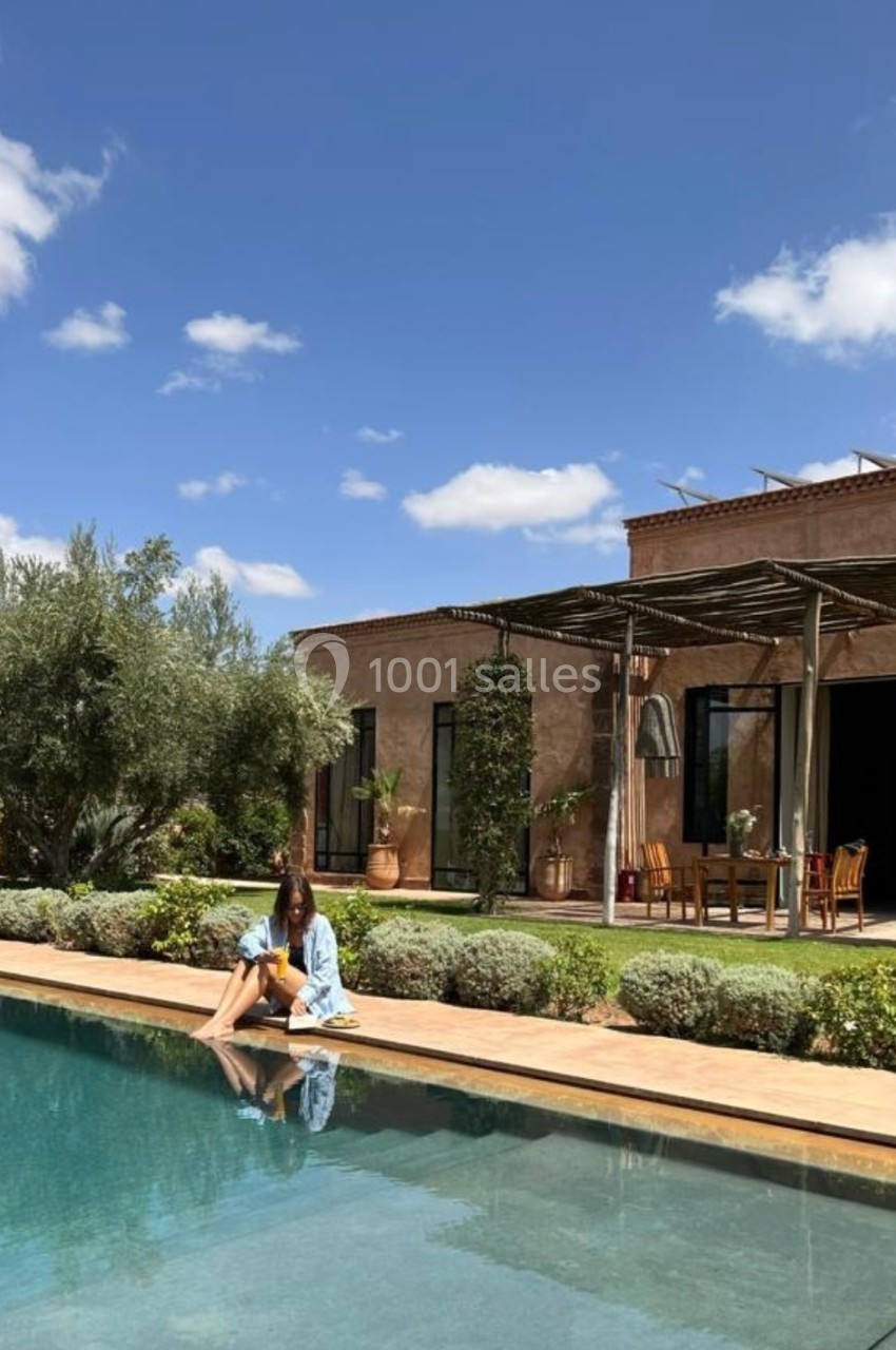 Femme assise au bord d'une piscine, devant une maison avec terrasse ombragée et jardin arboré sous un ciel bleu.