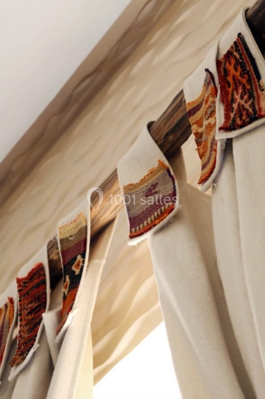 Rideau beige suspendu à une tringle en bois, décoré de bandes de tissu aux motifs colorés et géométriques.