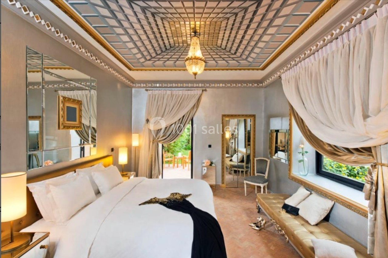 Chambre élégante avec lit double, décor doré, miroir mural, rideaux drapés et vue sur un espace extérieur verdoyant.