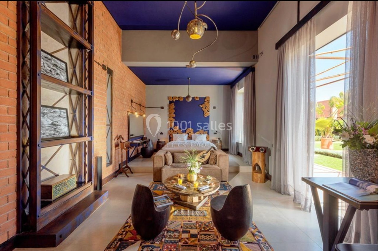 Chambre lumineuse avec murs en briques, plafond bleu, lit double, coin salon et grandes fenêtres donnant sur un jardin.