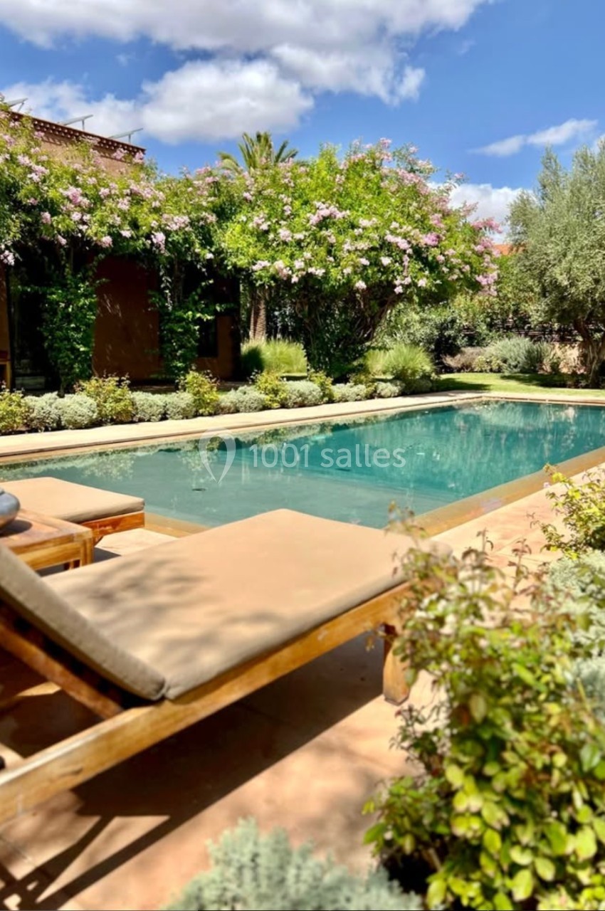Transat en bois près d'une piscine entourée de végétation luxuriante sous un ciel bleu avec quelques nuages.
