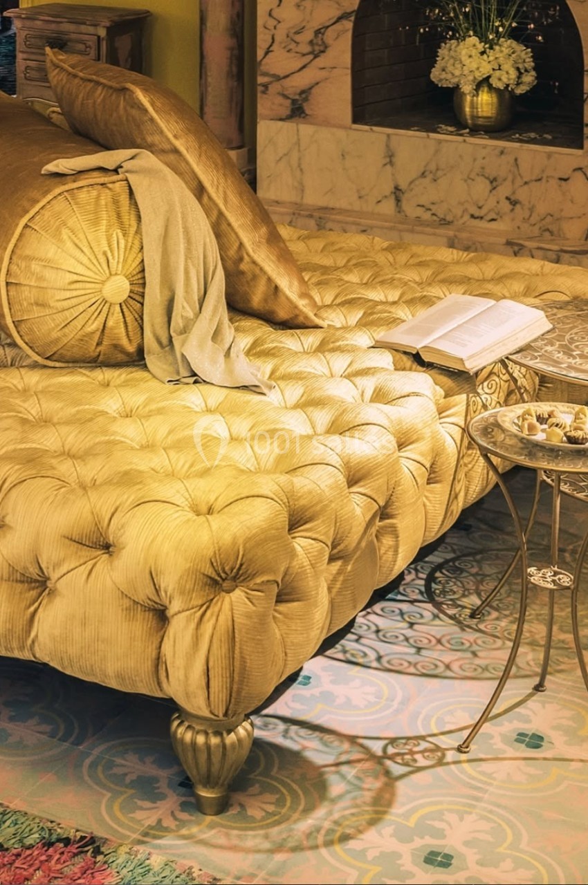 Lit capitonné doré avec coussins assortis, table d'appoint en métal, livre ouvert et cheminée en arrière-plan.