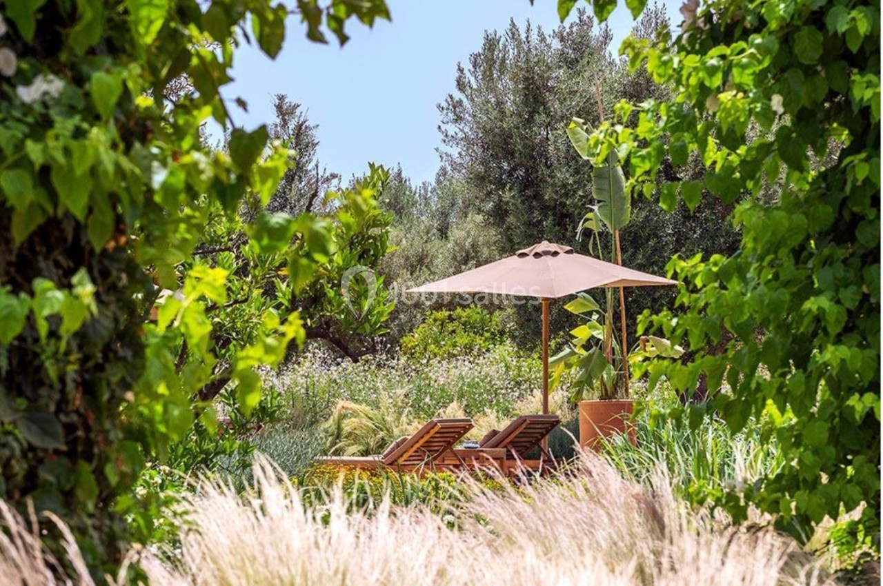 Deux chaises longues en bois sous un parasol beige, entourées de végétation luxuriante dans un jardin ensoleillé.