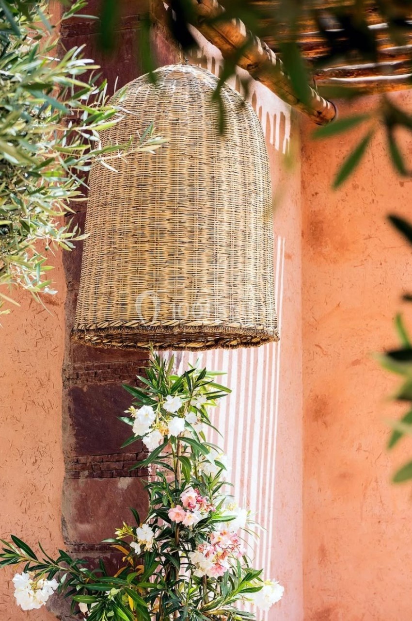 Suspension en osier tressé accrochée à un mur en stuc rose, entourée de plantes et de fleurs.