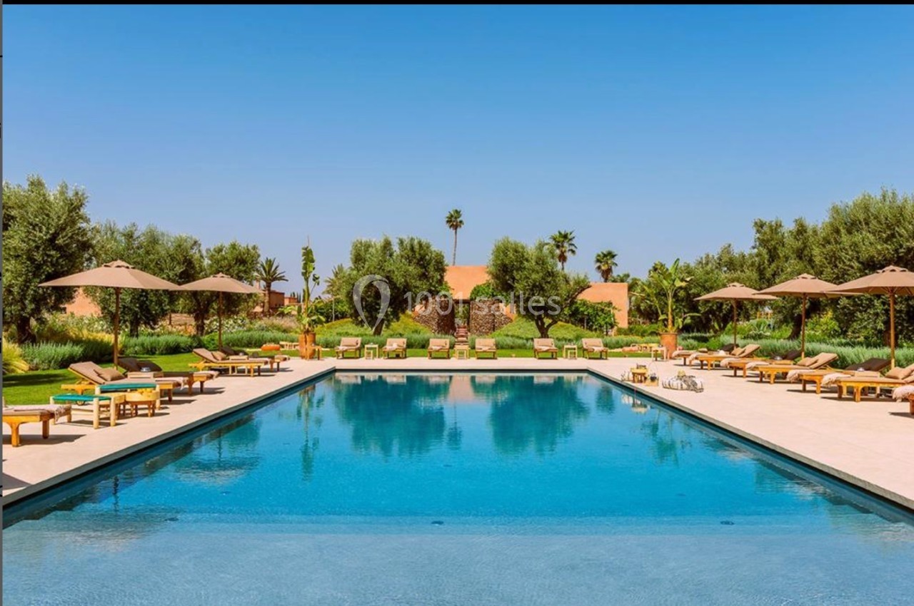 Piscine extérieure entourée de transats, parasols et végétation luxuriante sous un ciel bleu clair.