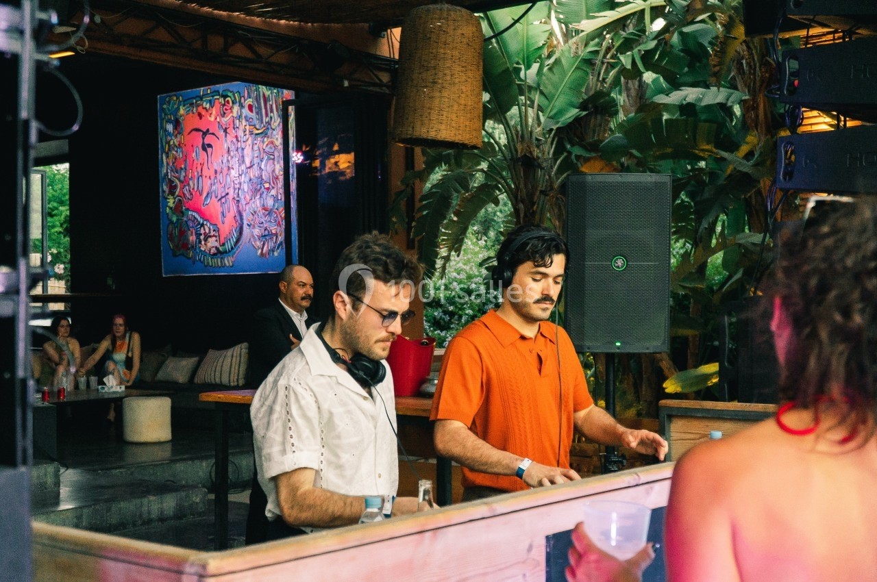 Deux DJ mixent de la musique dans un espace extérieur décoré de plantes et d'une peinture colorée en arrière-plan.