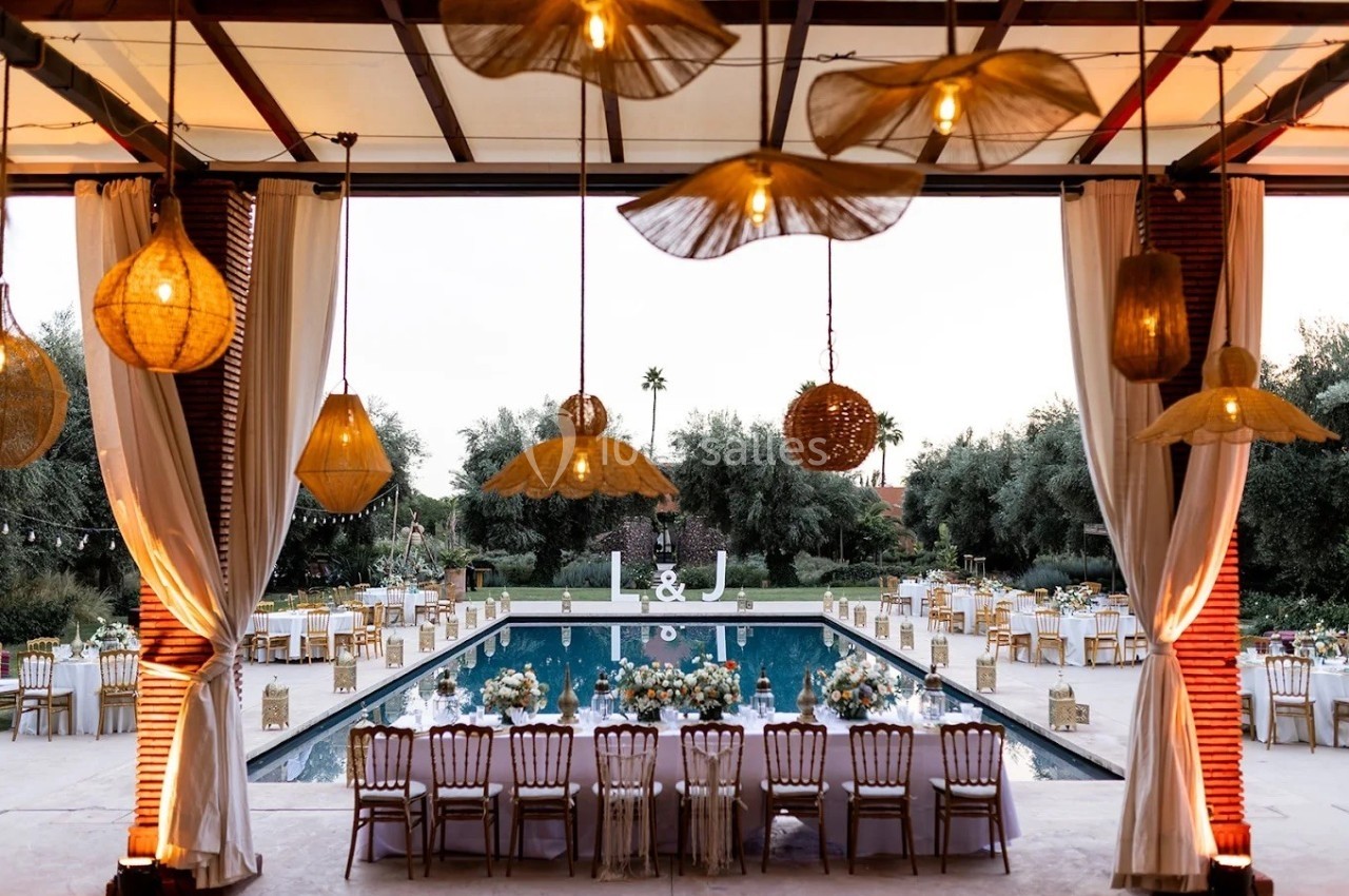 Vue d'une piscine entourée de tables décorées pour un événement, avec des lampes suspendues et un ciel dégagé en arrière…