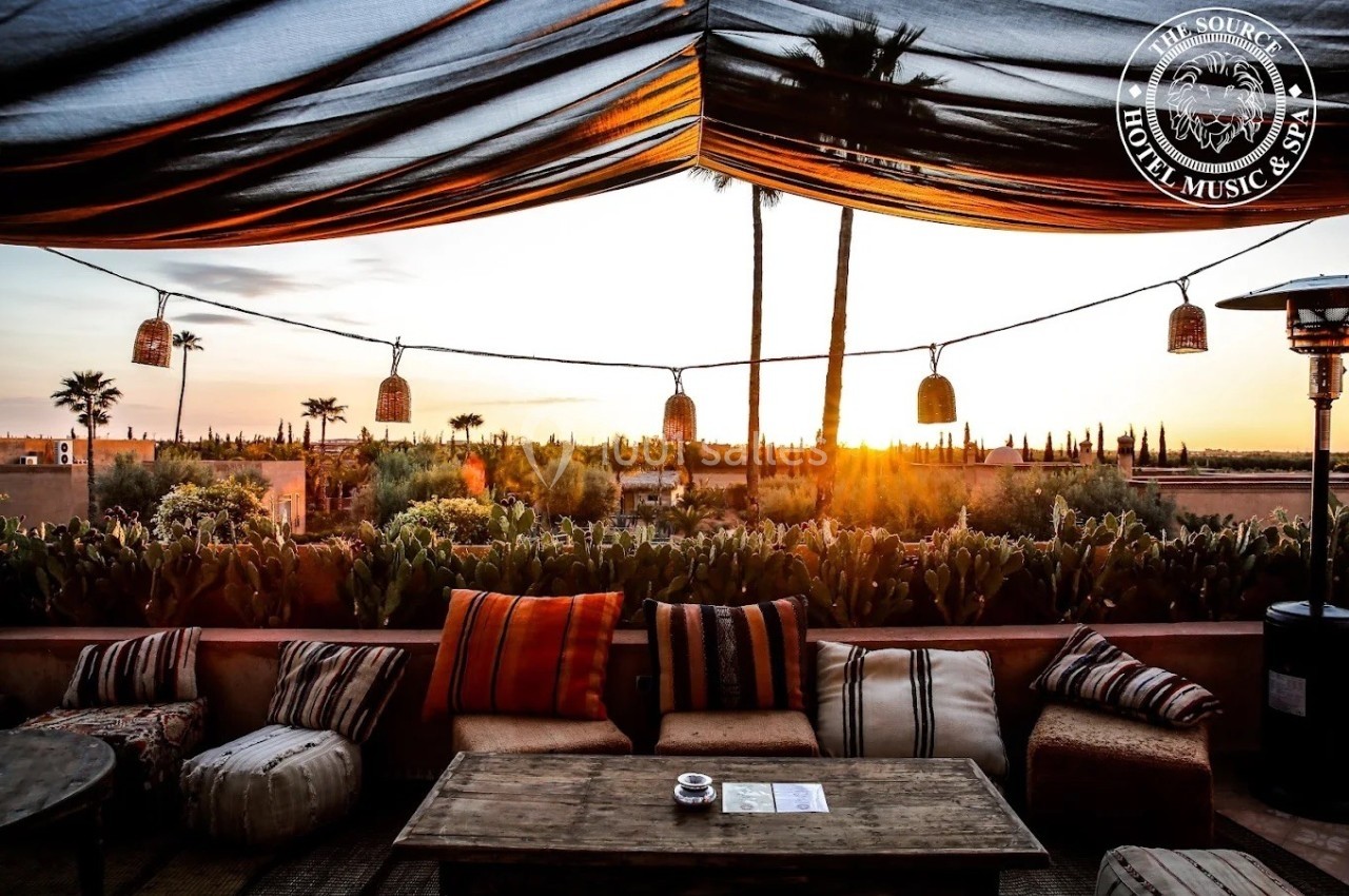 Terrasse avec mobilier en bois et coussins, vue sur un coucher de soleil, palmiers et végétation en arrière-plan.