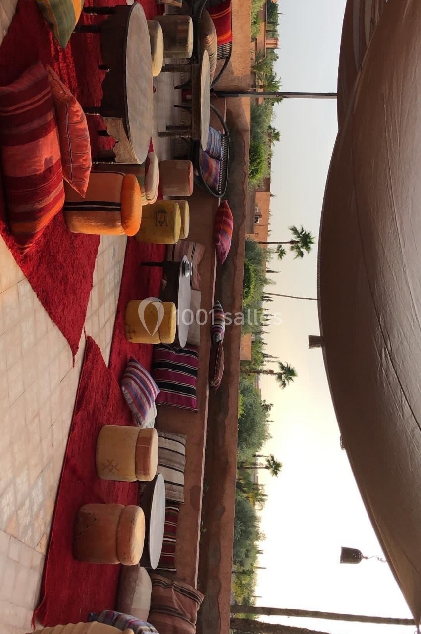 Espace extérieur avec tables basses, coussins colorés et tapis rouges, entouré de murs en terre cuite et végétation.