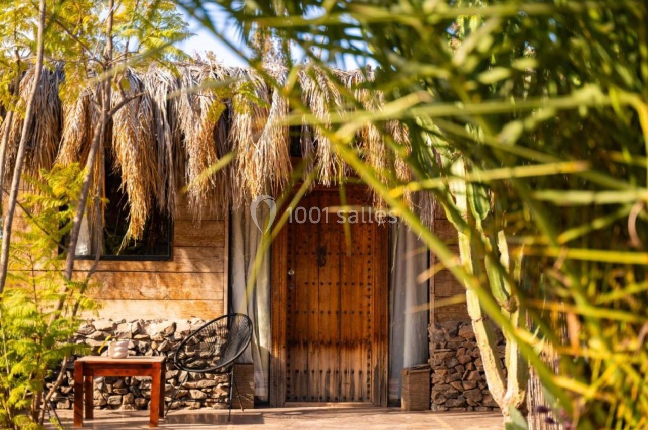 Entrée d'une maison en bois avec porte en bois sculpté, toit en chaume et végétation tropicale au premier plan.