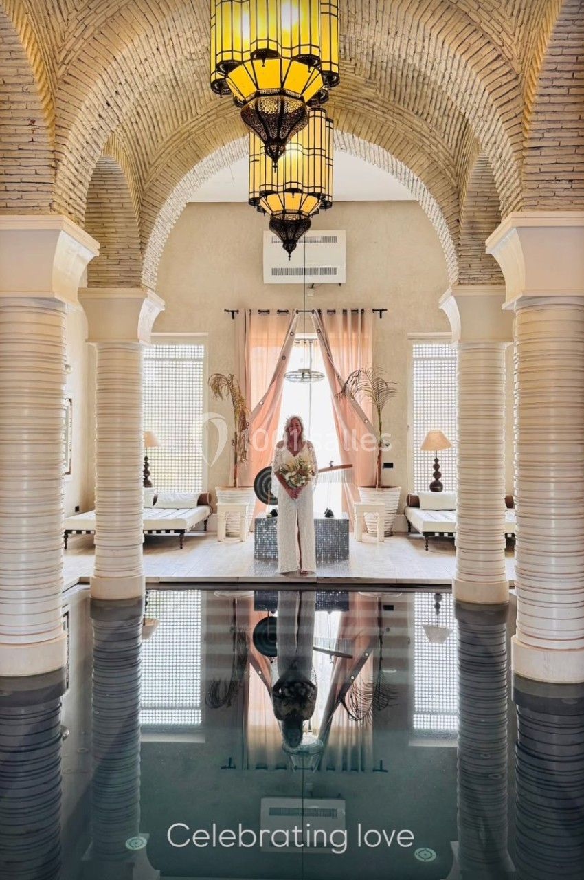 Une femme en robe blanche tient un bouquet dans une salle voûtée avec piscine intérieure et lumière naturelle.