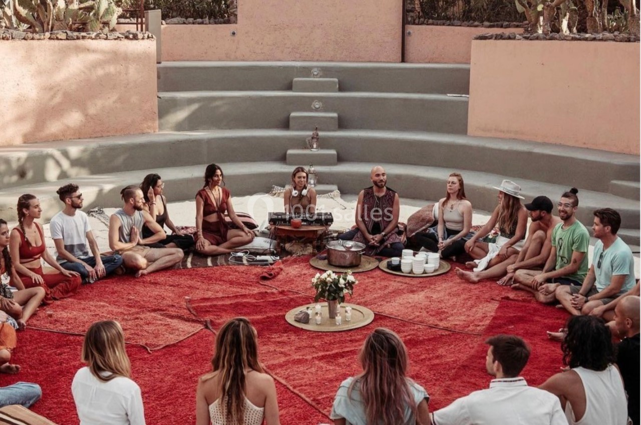 Un groupe de personnes assises en cercle sur des coussins autour d'un tapis rouge dans un espace extérieur.