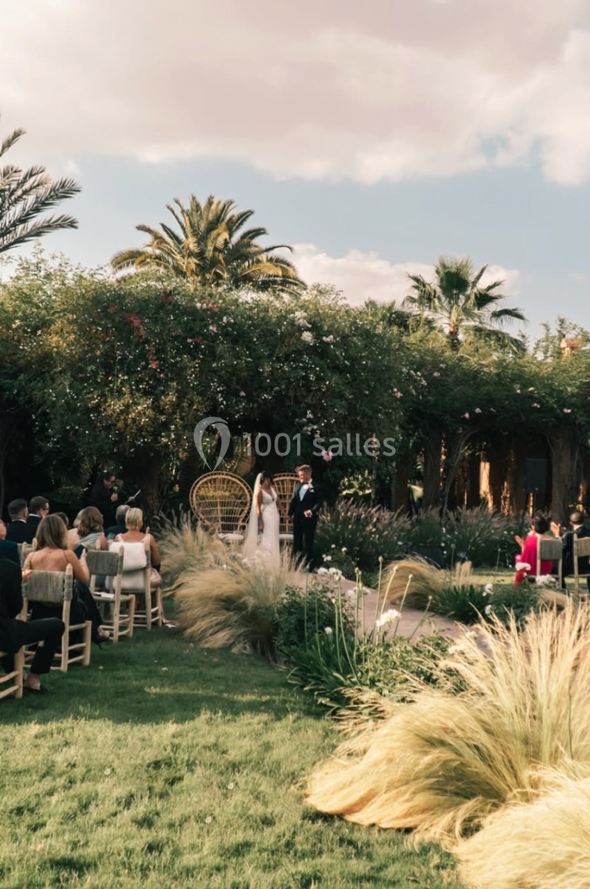 Cérémonie de mariage en extérieur avec des invités assis, un couple debout devant une arche végétale.