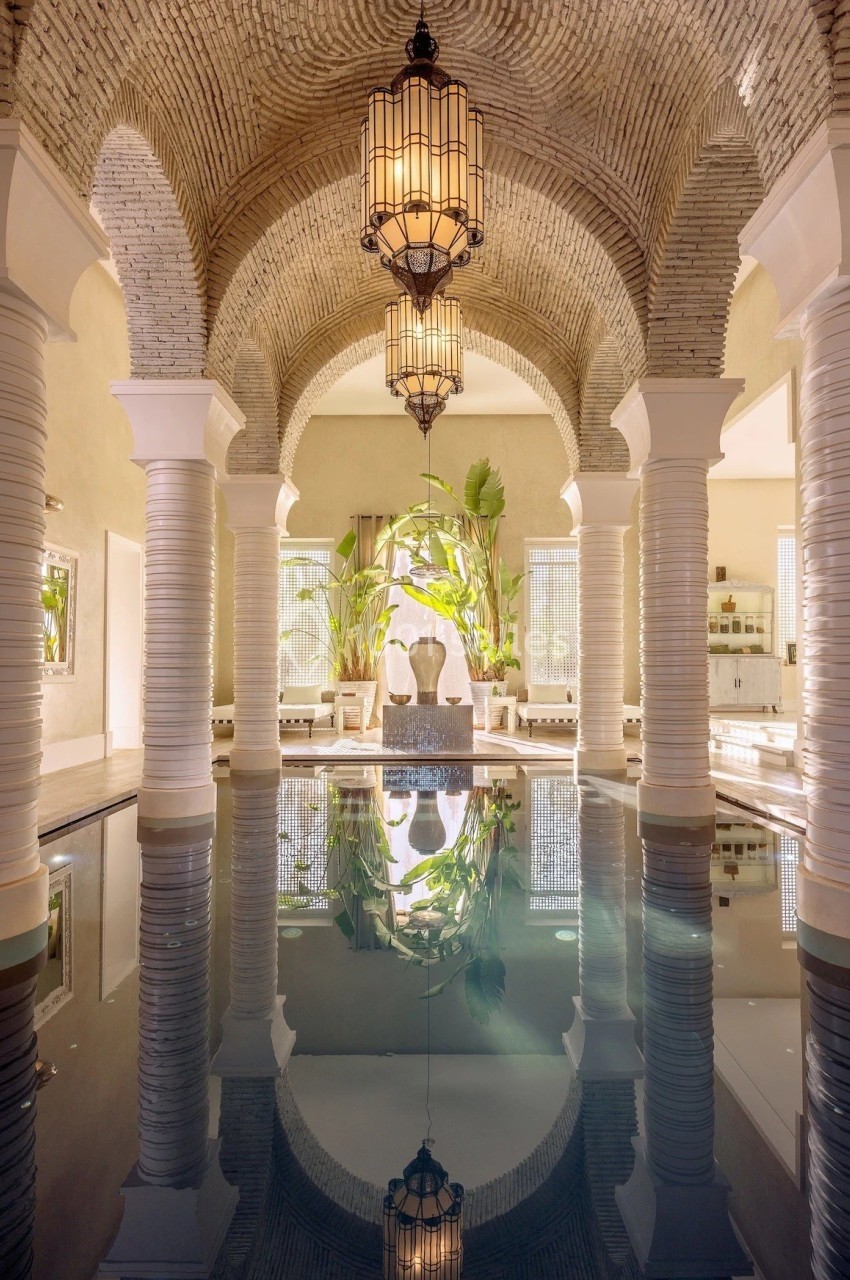 Piscine intérieure entourée d'arches en pierre, éclairée par des lanternes suspendues et décorée de plantes tropicales.