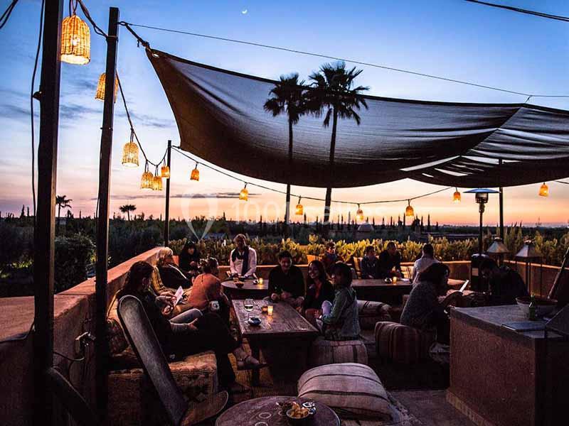Terrasse extérieure au coucher du soleil avec des lampes suspendues, des canapés et des personnes assises.